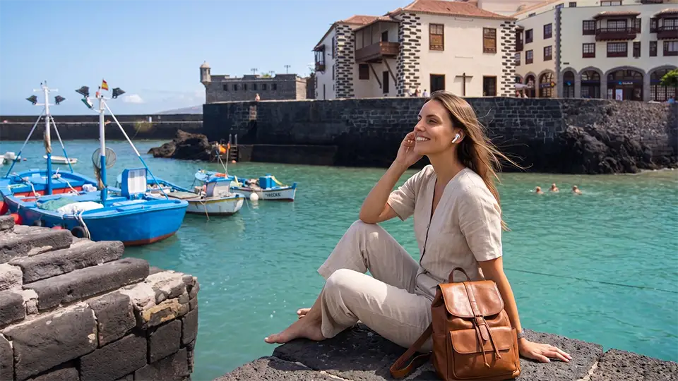 A young traveller sitting on the harbour breakwater, listening to an audio guide, gazing out at the sea and the colourful fishermen’s boats, in Puerto de la Cruz, Tenerife