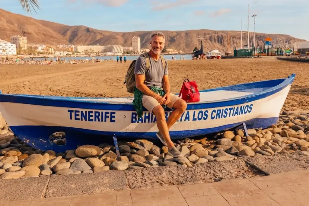 A traveller sitting in a boat beached on the sand, enjoying an immersive audio experience at Los Cristianos Beach, Costa Adeje, Tenerife