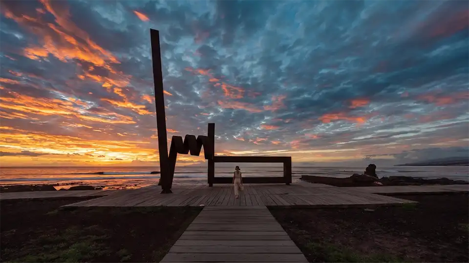 Eine Frau spaziert bei Sonnenuntergang zur Skulptur 'La Trampa del Viento', Costa Adeje, Teneriffa